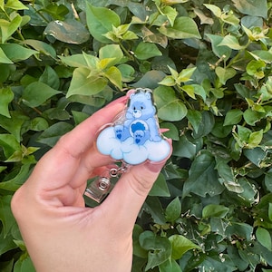 Może przedstawiać: Niebieska odznaka Care Bear z niebieskim misiem siedzącym na białej chmurze. Odznaka ma przezroczystą plastikową osłonę i srebrny metalowy klips. Tło to bujne zielone liście.