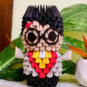 May include: A black, gray, red, and yellow origami figure of Harry Potter with round glasses. The figure is holding a wand in its right hand.
