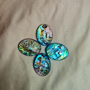 May include: Four oval-shaped abalone shell cabochons featuring a vibrant, iridescent surface with swirling patterns of deep blue, bright green, gold, and purple hues. Each piece displays a polished, reflective finish with unique natural textures.