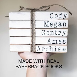 May include: A stack of five white books with names written on the spines: Cody, Megan, Gentry, Ames, and Archie with a paw print. The books are tied together with twine and the text "MADE WITH REAL PAPERBACK BOOKS" is printed below the stack.