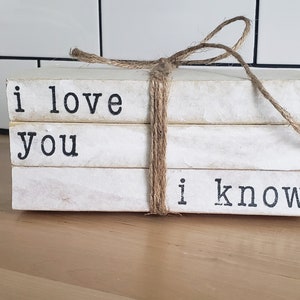 May include: A stack of three white books with black lettering. The books are tied together with twine and the text on the books reads "i love you i know".