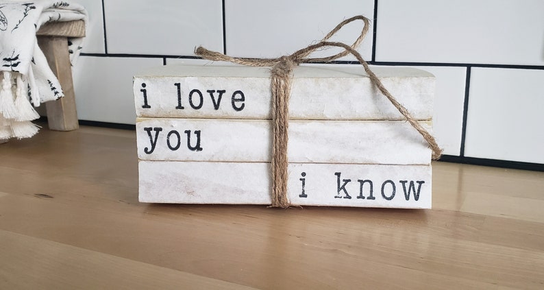 May include: A stack of three white books with black lettering. The books are tied together with twine and the text on the books reads "i love you i know".