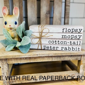 May include: A set of four white books stacked on top of each other. The books are tied together with twine. The books have the words "flopsy", "mopsy", "cotton-tail", and "peter rabbit" written on them. The books are sitting on a wooden table with a ceramic bunny figurine and green plants.
