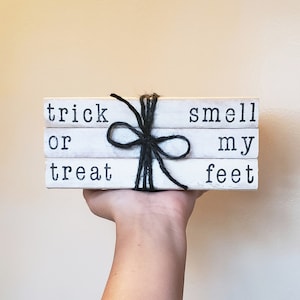 May include: Three white wooden blocks stacked and tied with black twine. The top block reads "trick", the middle block reads "or", and the bottom block reads "treat". The right side of the blocks reads "smell", "my", and "feet".