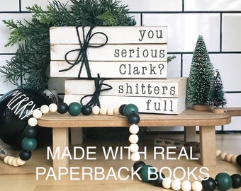 May include: A Christmas-themed display featuring stacked white paperback books with the phrases "you serious Clark?" and "shitters full" printed in black. A black and white beaded garland and a black ornament with "MERRY" in white complete the festive arrangement.
