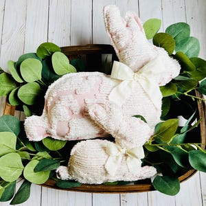 May include: Two plush pink and white bunny decorations with cream-colored bows, nestled in a wooden basket surrounded by green foliage. The bunnies have a textured, patterned design. A charming Easter or spring-themed decorative item.