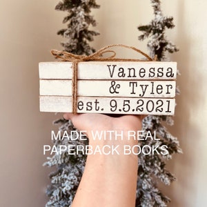 May include: A stack of three white paperback books with the text "Vanessa & Tyler est. 9.5.2021" printed on the top book. The books are tied together with twine and the text "MADE WITH REAL PAPERBACK BOOKS" is printed below the books.