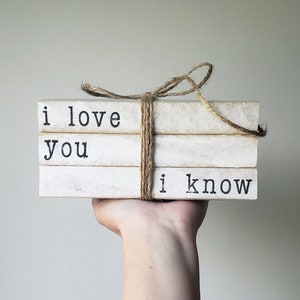 May include: A stack of three white books with black text. The books are tied together with twine and the text reads "i love you i know".