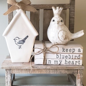 Puede incluir: Una casita de p&aacute;jaros de cer&aacute;mica blanca con un lazo de arpillera marr&oacute;n se sienta en un banco de madera r&uacute;stico con una figurita de p&aacute;jaro de cer&aacute;mica blanca que lleva una corona. El banco tiene una pila de tres libros blancos con el texto "keep a bluebird in my heart" impreso en la portada.