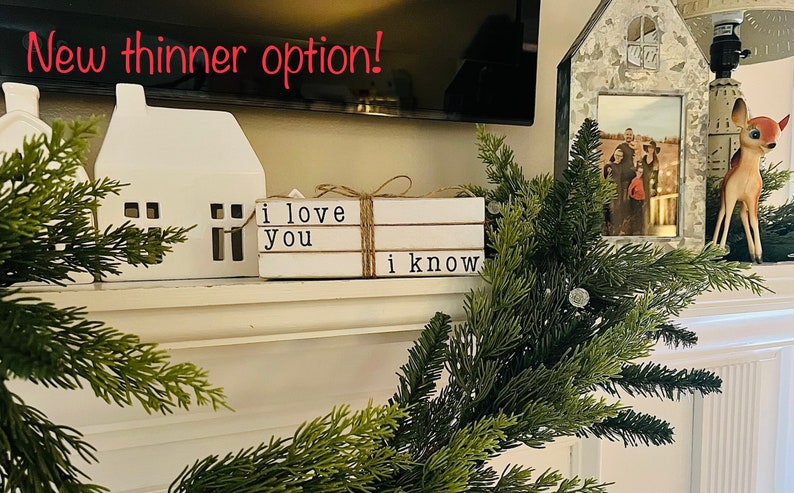 May include: White ceramic houses with window cutouts, a stack of three white books with the text "i love you" and "i know" on them, a metal house with a photo inside, and a green leafy garland with a white deer figurine.