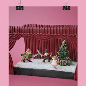 May include: A miniature stage set with a Christmas theme. The scene includes a Christmas tree, presents, and figurines of characters in a play. The backdrop features red curtains and a pink background.