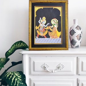 May include: A framed painting featuring two figures with blue and green skin tones, dressed in vibrant yellow, red, and orange traditional attire, dancing on a checkered floor against a black background, enclosed in a gold-toned rectangular frame.