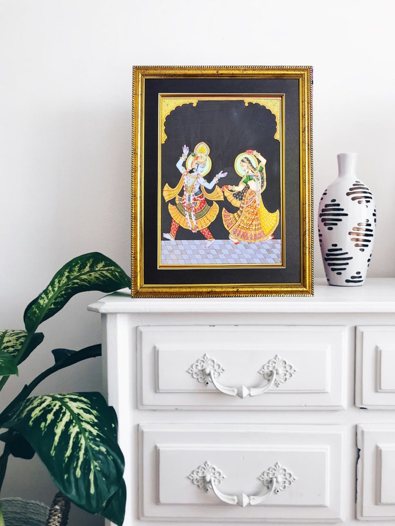May include: A framed painting featuring two figures with blue and green skin tones, dressed in vibrant yellow, red, and orange traditional attire, dancing on a checkered floor against a black background, enclosed in a gold-toned rectangular frame.
