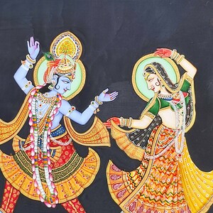 May include: A painting on a black background depicting two figures dancing. The figure on the left has blue skin, wears a crown with a peacock feather, and is adorned with jewelry and a floral garland. The figure on the right wears a green and yellow patterned dress with jewelry.