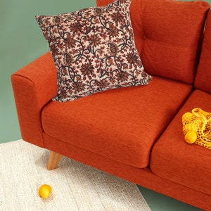 May include: A burnt orange sofa with a floral patterned pillow and a yellow mesh bag with lemons on the seat. The sofa is on a white rug with two lemons on the floor.