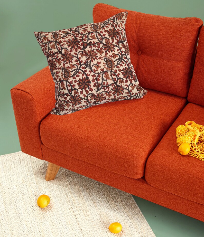 May include: A burnt orange sofa with a floral patterned pillow and a yellow mesh bag with lemons on the seat. The sofa is on a white rug with two lemons on the floor.