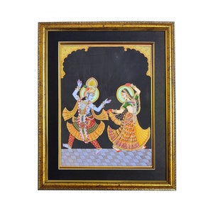 May include: A framed painting on a black background depicting two figures in traditional attire dancing on a checkered floor. The figure on the left has blue skin and wears a yellow and red outfit, while the figure on the right wears a green and yellow dress. Both figures have halos.