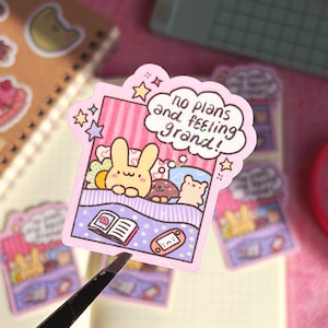 Op de afbeelding: Een roze sticker met een cartoon illustratie van een konijn, een hond en een beer in bed, met de tekst "no plans and feeling grand!" in een wolk. De sticker is versierd met sterren en wordt vastgehouden door een pincet.