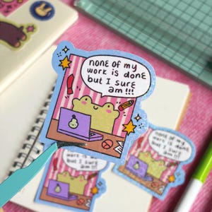 May include: A sticker featuring a cartoon frog at a desk with a laptop and pencil, with the text "none of my work is done but I sure am!!!". The sticker has a blue border and star accents. Other stickers and a notebook are in the background.