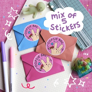 May include: A collection of stationery items including three folded paper envelopes in blue, pink, and peach. Each envelope has a circular sticker with a bunny design and the text "OOPSIE BAG". The image also shows markers, a paper cutter, and small figurines. Text reads "mix of 5 Stickers".