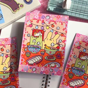 May include: A rectangular art print featuring a colourful cartoon illustration of a yellow rabbit and a green frog eating at a table. The scene includes bowls of noodles, dumplings, and sushi, decorated with a border of pink stars and string lights.