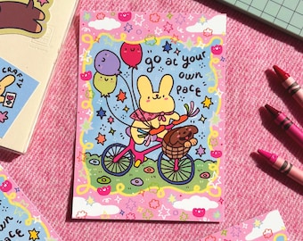 May include: A colorful illustration features a yellow bunny riding a pink bicycle, holding balloons and a basket with a brown animal. The text "go at your own pace" is written on the image. The artwork is surrounded by a pink border with stars and clouds.