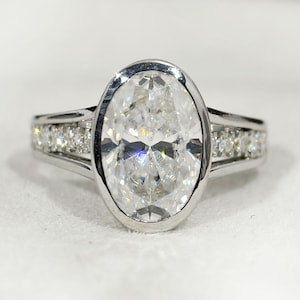 Pode incluir: Um anel de prata com um grande diamante de corte oval como peça central. O diamante está engastado em uma moldura e é ladeado por diamantes menores na banda. O anel é exibido em uma superfície branca.