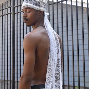 Könnte beinhalten: Weiße Durag aus Spitze mit floralem Muster. Das Kopftuch ist hinten gebunden und hat einen langen, fließenden Schwanz. Die Kopfbedeckung wird von einer Person in hellblauen Jeans getragen.