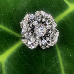 May include: A silver-toned flower-shaped brooch with clear, sparkling rhinestones. The brooch features a central, large rhinestone surrounded by smaller stones and intricate filigree detailing. The brooch is set against a dark green leaf.