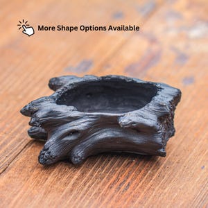 May include: A black planter shaped like a piece of weathered driftwood with a hollow centre. The surface features detailed wood grain textures and knots. Above the planter, the text reads: More Shape Options Available with a hand icon.