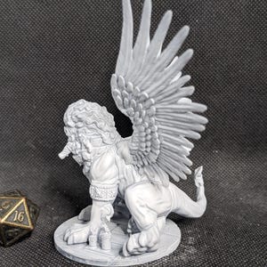 May include: A light gray unpainted resin miniature of a winged lion-like creature with a textured mane and detailed feathers, standing on a circular base next to a twenty-sided die with gold-colored numbers on a dark gray surface.