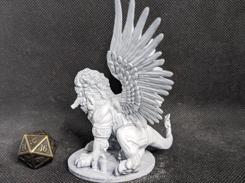 May include: A light gray unpainted resin miniature of a winged lion-like creature with a textured mane and detailed feathers, standing on a circular base next to a twenty-sided die with gold-colored numbers on a dark gray surface.
