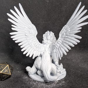 May include: A light gray unpainted miniature figurine of a winged lion-like creature with a long tail, standing on a circular base. Beside the figurine is a dark metallic twenty-sided die with gold-colored numbers showing 14 and 16.