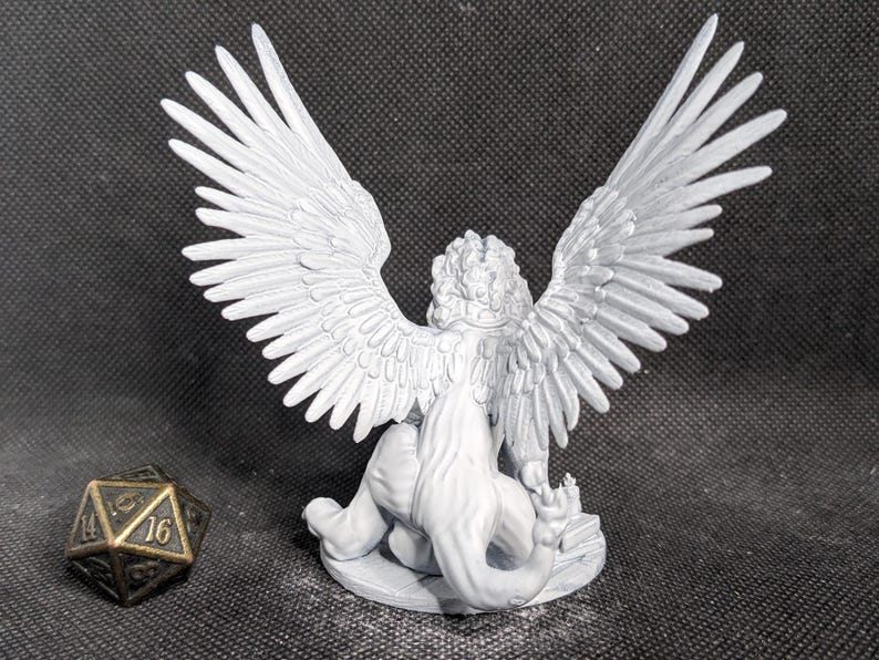 May include: A light gray unpainted miniature figurine of a winged lion-like creature with a long tail, standing on a circular base. Beside the figurine is a dark metallic twenty-sided die with gold-colored numbers showing 14 and 16.