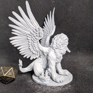 May include: A light gray unpainted resin miniature of a winged lion creature with a thick mane, standing on a circular base with small books. Beside the creature sits a dark metallic twenty-sided die with gold-colored numbers visible.