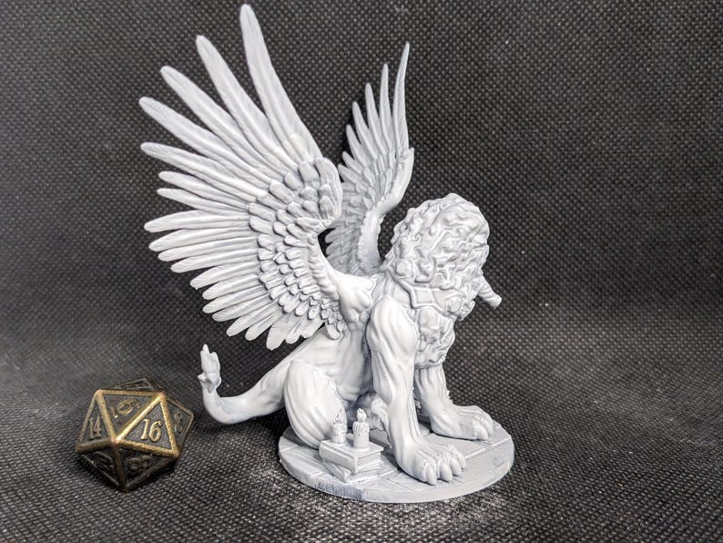May include: A light gray unpainted resin miniature of a winged lion creature with a thick mane, standing on a circular base with small books. Beside the creature sits a dark metallic twenty-sided die with gold-colored numbers visible.