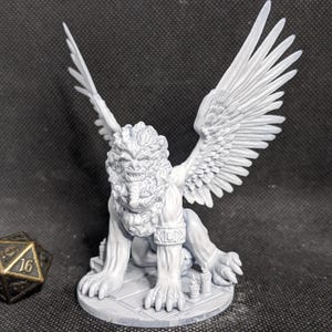 May include: A light gray, unpainted resin miniature of a winged lion creature with a textured mane, standing on a circular base with small candle details. Beside the figure sits a twenty-sided die with gold-colored numbers on a dark surface.