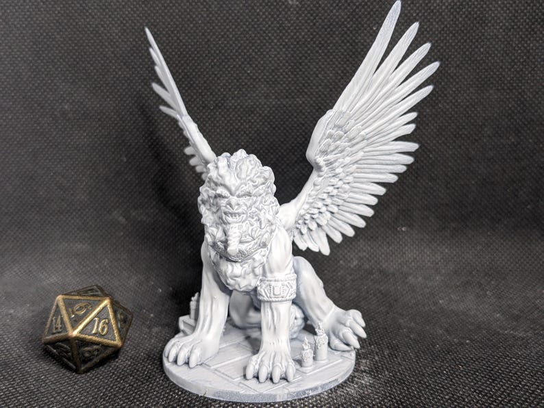 May include: A light gray, unpainted resin miniature of a winged lion creature with a textured mane, standing on a circular base with small candle details. Beside the figure sits a twenty-sided die with gold-colored numbers on a dark surface.