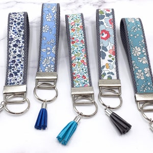 May include: Five keychains with different floral patterns on the fabric strap. Each keychain has a silver ring and a tassel. The tassels are blue, turquoise, and gray.