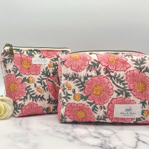 May include: Two floral print cosmetic bags. The bags feature a white background with a pattern of pink flowers, green leaves, and yellow accents. The bags have a zipper closure and a small tag that reads "Helen K. Studio".