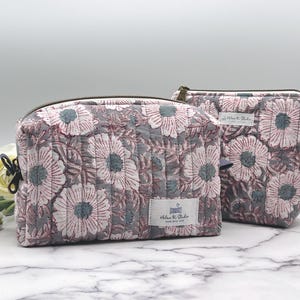 May include: Two floral print zippered cosmetic bags. The larger bag is grey with pink and blue flowers. The smaller bag is white with blue and pink flowers. Both bags have the text "Helen & Blake" and "Made with Love" printed on them.