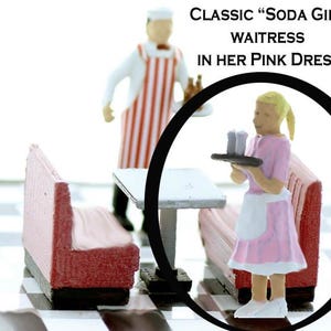 May include: A miniature figurine of a waitress wearing a pink dress and white apron, holding a tray with two glasses. The figurine is standing in front of a miniature diner booth and table. The text "CLASSIC "SODA GIRL" WAITRESS IN HER PINK DRESS" is visible above the figurine.