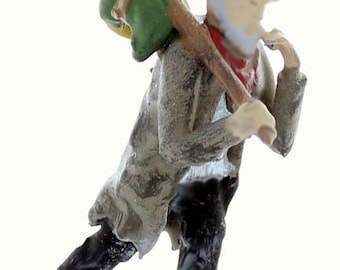 May include: A figurine of a person wearing a brown hat and a gray coat, carrying a green bag on their back and a wooden stick in their hand.
