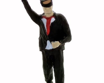 May include: A black and red painted miniature figure of a train conductor wearing a black cap and a red tie, with his arm raised in a signaling gesture.