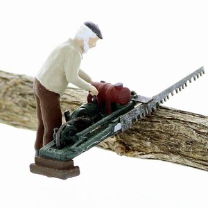 May include: A miniature figurine of a person operating a small, green and red chainsaw on a wooden log. The chainsaw has a metal blade with teeth.