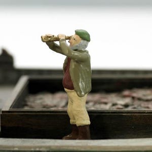 May include: A miniature figurine of a person wearing a green hat and a brown jacket, holding a telescope and looking out into the distance.