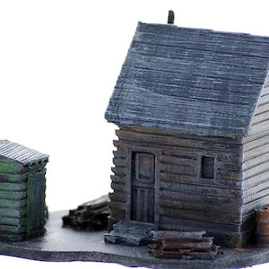 May include: A miniature model of a rustic wooden cabin with a weathered gray roof and a green outhouse. The cabin has a door and a window, and the outhouse has a door. The model is set on a base with a few pieces of wood and some barrels.