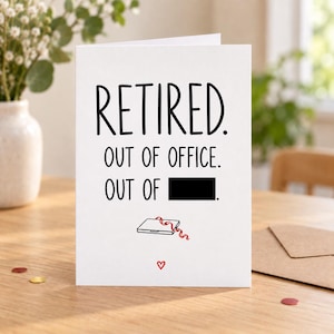 Lifestyle mock-up of a standing retirement card with an out-of-office joke, one word covered by a black bar, and a laptop doodle. On a wooden table with a kraft envelope to the right and red/gold confetti.