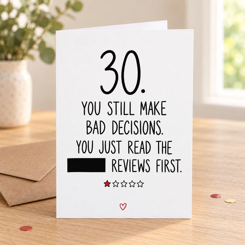 Lifestyle mock-up of a standing 30th birthday card about bad decisions and reviews, with one word covered by a black bar and a one-star doodle. On a wooden table beside a Kraft envelope and red/gold confetti.