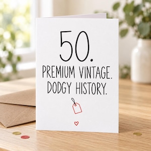Lifestyle mock-up of a standing card reading “50. PREMIUM VINTAGE. DODGY HISTORY.” with a small red auction tag. On a wooden table beside a Kraft envelope and red/gold confetti.